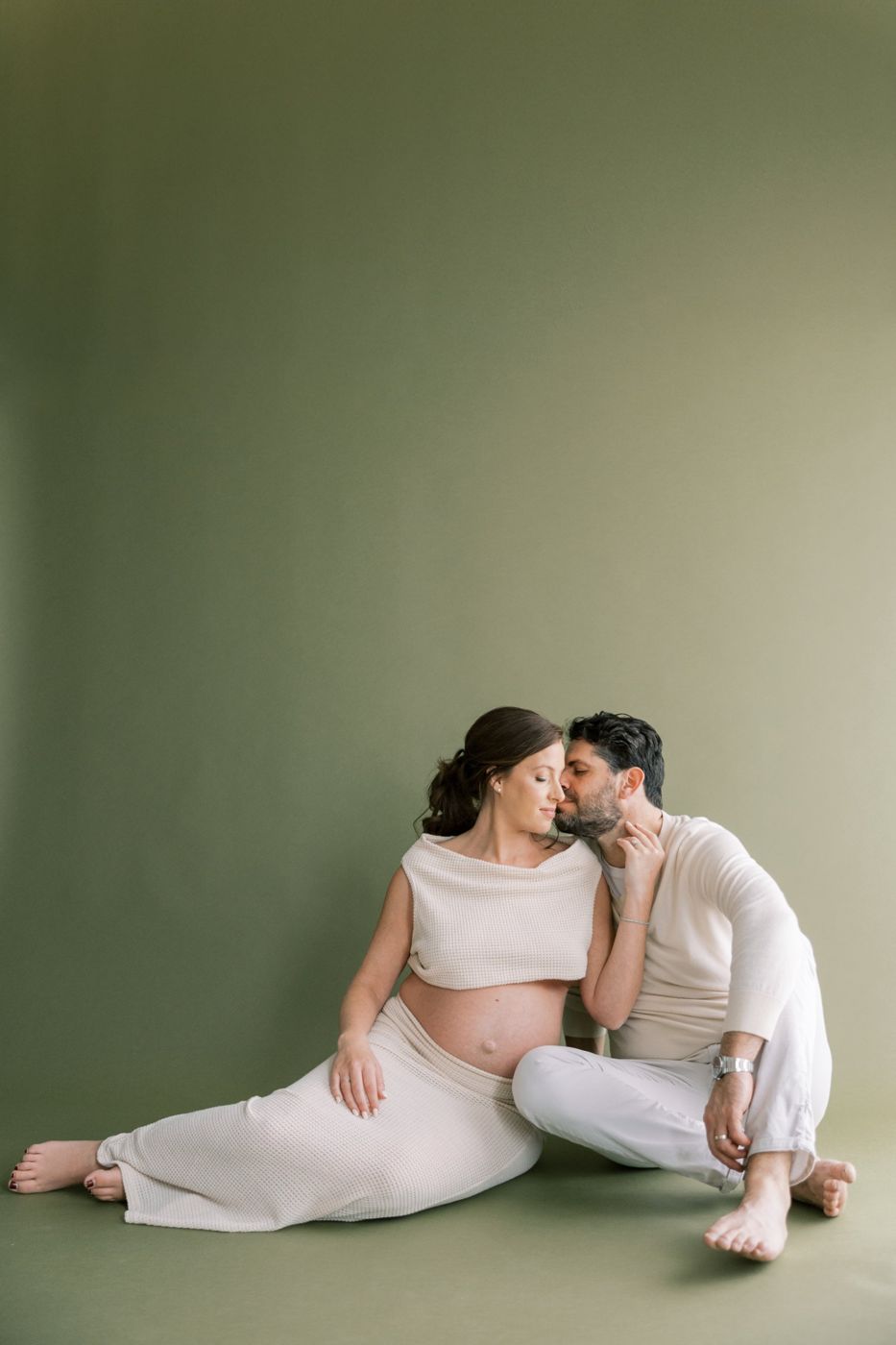 Expecting couple sitting close on the floor against a sage green backdrop