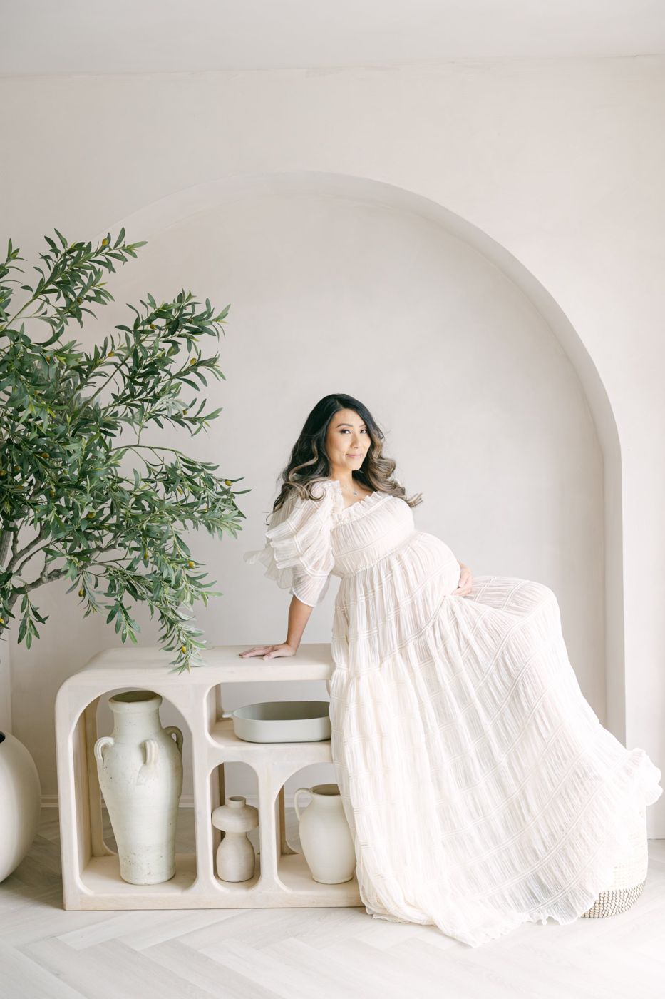 Expecting mother in a flowing white gown leaning on a console under the arch beside the olive tree