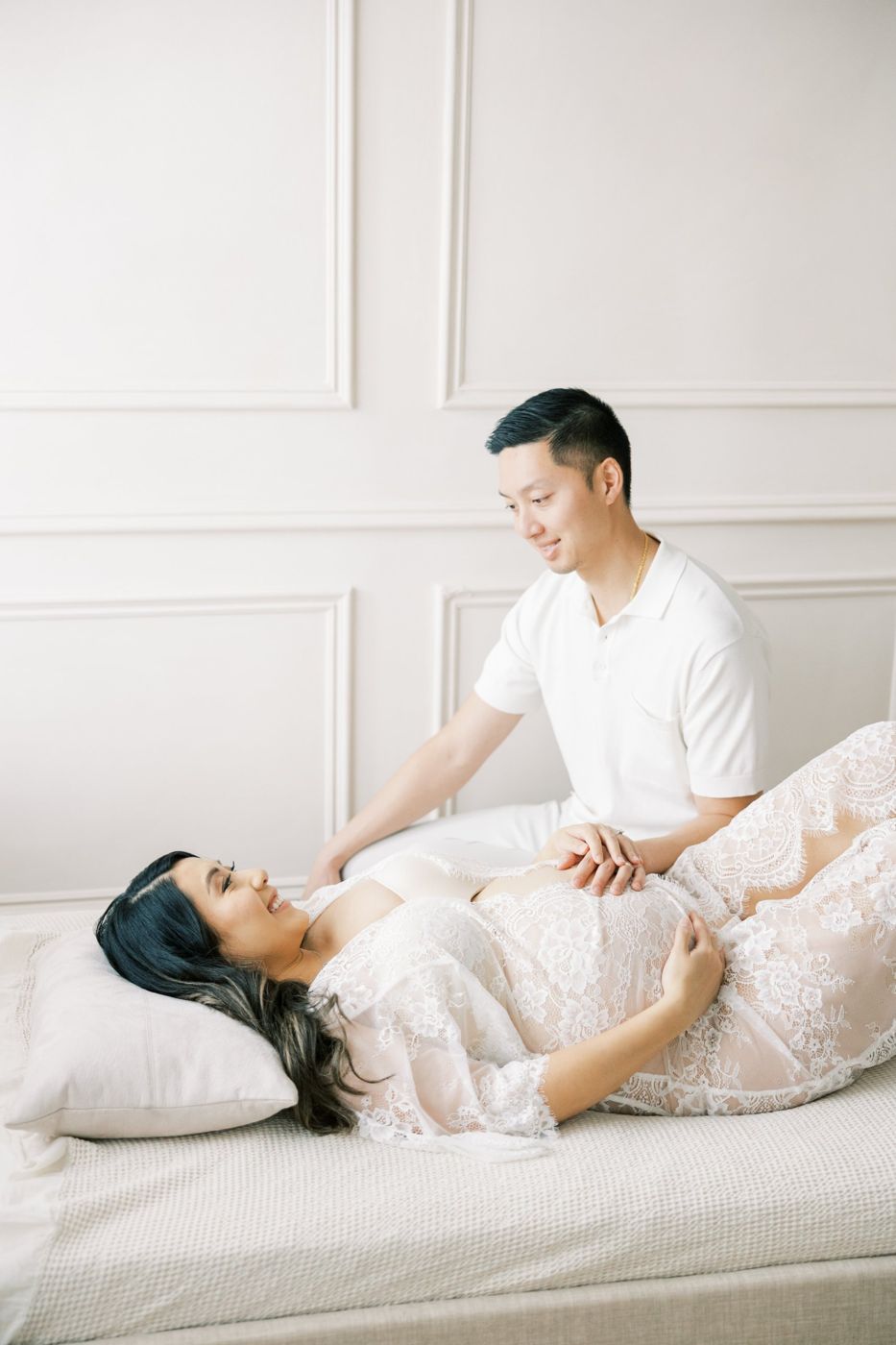 Expecting mother in white lace on the daybed with her partner, molding wall behind