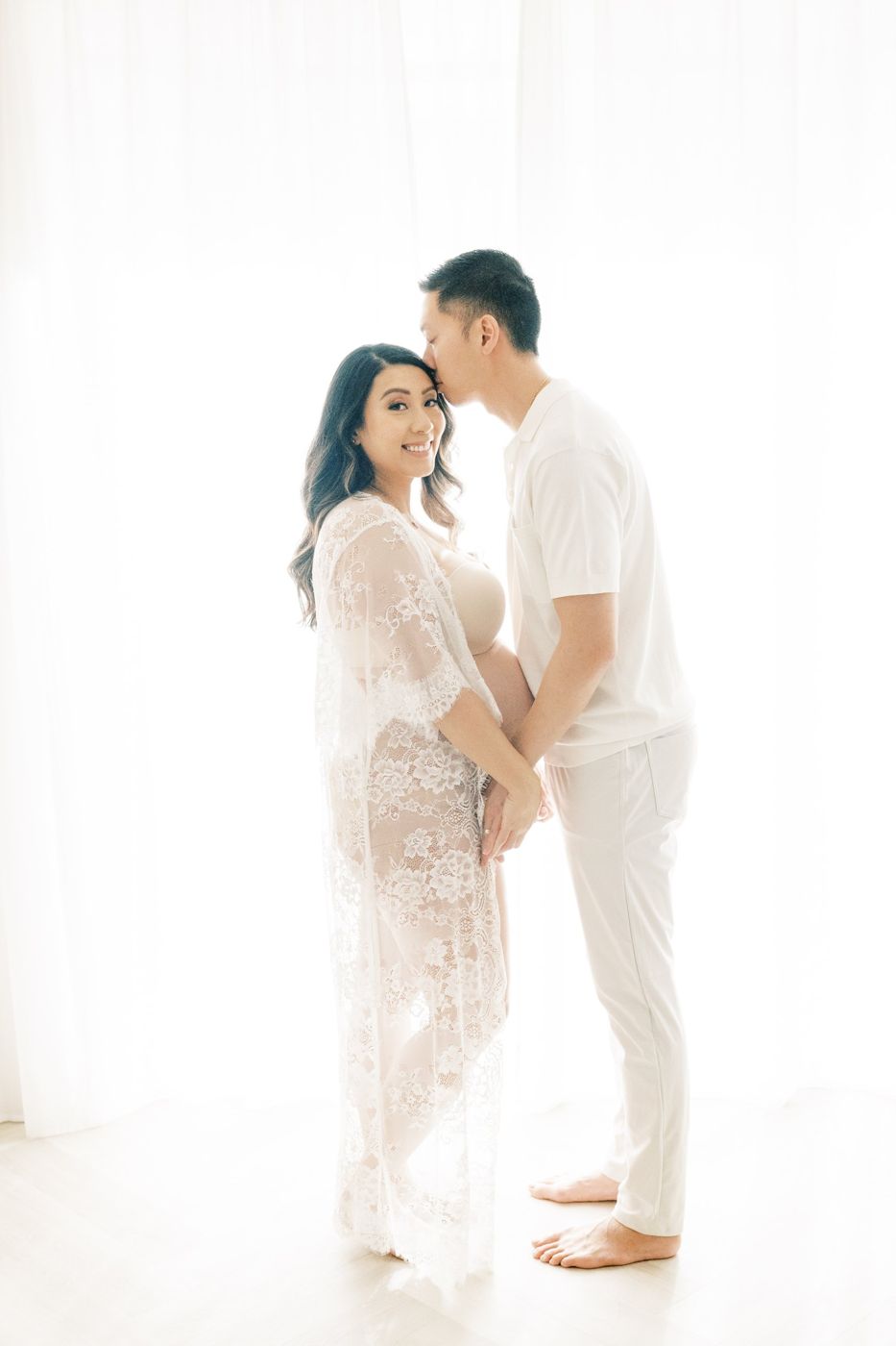 Couple in white backlit by sheer-curtained windows, partner kissing her forehead
