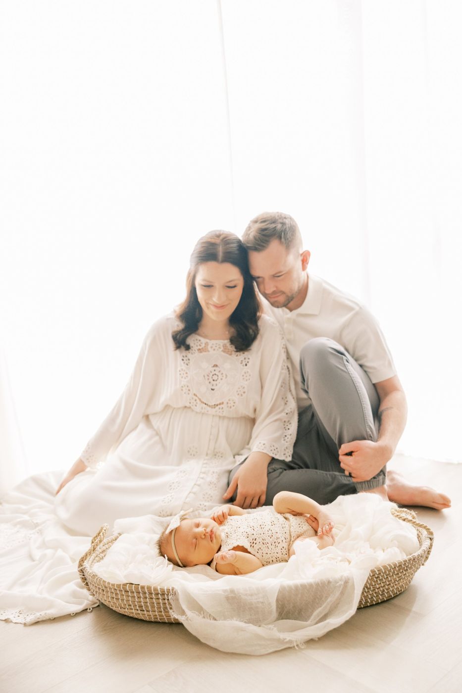 New parents sitting on the floor in window light, smiling at their sleeping newborn in a woven basket