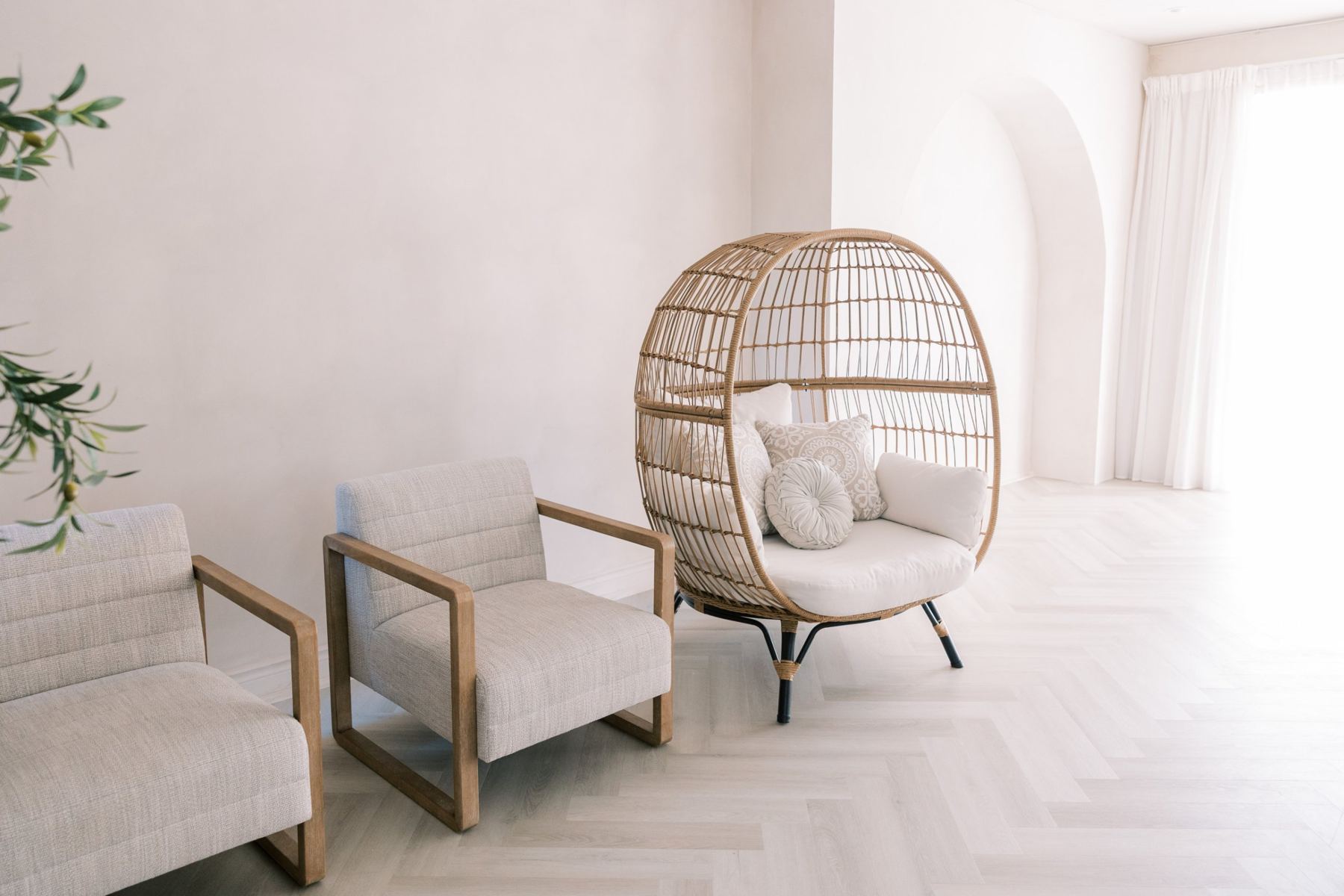Two wood-framed linen lounge chairs and a rattan egg chair on pale herringbone floors, with the arch and bright windows behind